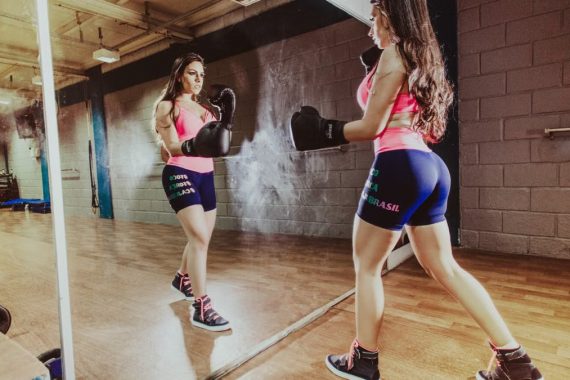 Back view concentrated sportswoman with perfect body wearing sportswear and boxing gloves punching air while looking in big wall mirror in modern gym